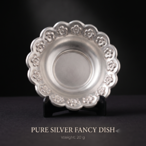 Pure silver fancy dish (20g) featuring intricate chased floral embossed patterns on a ruffled, scalloped rim, resting on a matte black display stand in a dark luxury studio. Excellent for home decor, gifting, or puja
